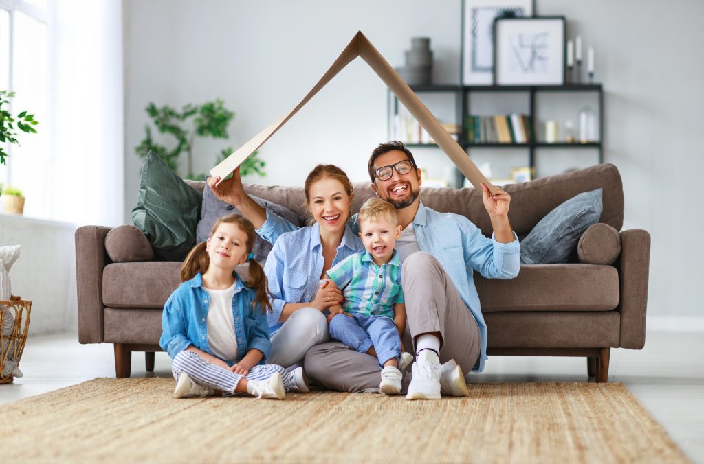 A family of four sitting in their living room holding a make-shift "roof" over their heads from cardboard.